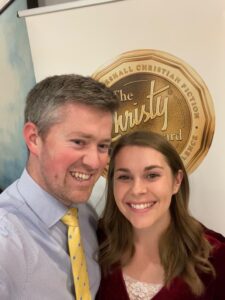 A smiling couple poses for a selfie in front of a banner featuring “The Christy Award” emblem. The man wears a light blue shirt with a yellow tie, and the woman wears a red velvet top with a lace neckline.