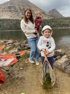 A smiling woman stands beside a mountain lake holding a young boy, while an older boy proudly shows a fishing net containing a small fish and green lake weeds. Camping chairs and fishing poles are visible near the rocky shoreline, with tall pine trees and mountains in the background.