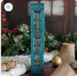A tall rectangular advent calendar box designed to look like a vintage teal bookshelf filled with colorful books, displayed among Christmas greenery and cozy holiday decor.