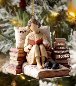 A personalized Christmas ornament depicting a woman named “Emily” sitting on a stack of books with a cup of tea, reading a book beside a small black cat, and surrounded by Christmas tree branches.