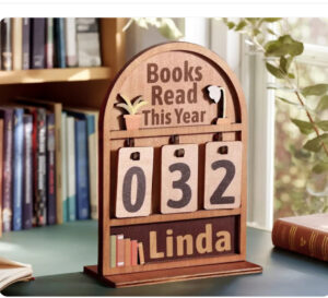 A wooden bookshelf-shaped display sign with the words “Books Read This Year” engraved at the top and hanging number cards showing 032. The bottom features a customizable nameplate reading “Linda” with small book illustrations.