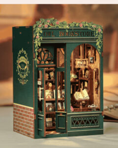 A detailed miniature model of a charming green storefront labeled “Owl Bookstore,” decorated with vines and pink roses around the sign. The tiny shop features glowing interior lights, shelves of miniature books, framed art, and a small open book on display, creating a cozy, magical bookstore scene — a perfect reader Christmas gift idea for book lovers and collectors.