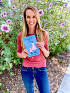 A woman standing outdoors in front of blooming pink and purple flowers, smiling while holding a copy of the book Beneath the Seams. She wears a coral top and jeans, and the book cover shows two dress forms with pink patterned dresses.