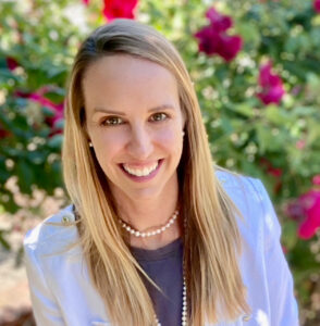 A woman smiling outdoors in front of vibrant pink and red flowers. She has long blonde hair and is wearing a light-colored jacket, a dark top, and a pearl necklace.