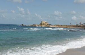 View of the coastline at Caesarea Maritima, showing turquoise Mediterranean waves rolling onto a sandy beach with ancient stone ruins and a fortress-like structure in the distance under a partly cloudy sky.