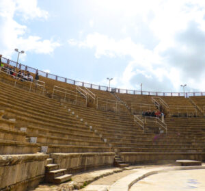 Stone seating inside the ancient Roman theater at Caesarea Maritima, showing curved rows of weathered steps, railings added for safety, and a small group of visitors sitting near the top under a bright, partly cloudy sky.