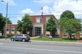 Red-brick county courthouse with white columns, mature trees, and an American flag, situated along a small-town street on a clear day.
