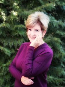 Author Linda Dindzans smiling warmly, wearing a plum sweater and posing with her hand resting on her cheek, standing in front of lush green evergreens