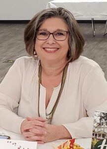 Author Regina Rudd Merrick smiling at a table, wearing glasses and a light-colored blouse, with short gray-brown hair in an indoor setting.