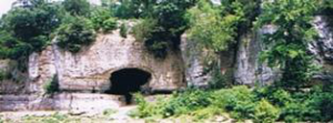 Large natural cave opening carved into a rocky limestone cliff, surrounded by green trees and vegetation along a riverbank.
