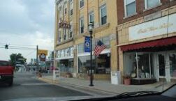 Small-town downtown street with historic brick storefronts, local shops, streetlamps, and an American flag, viewed from inside a car in a quiet rural community.