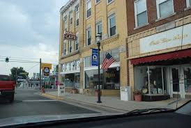Small-town downtown street with historic brick storefronts, local shops, streetlamps, and an American flag, viewed from inside a car in a quiet rural community.