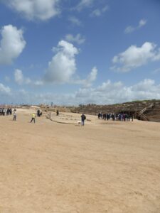 Visitors walking through the sandy arena of the ancient hippodrome at Caesarea Maritima, with stone seating and ruins in the background under a bright sky filled with scattered clouds