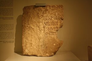 Photograph of the Pilate Stone, a weathered limestone block with a partial Latin inscription referencing Pontius Pilate, displayed upright in a museum under warm lighting