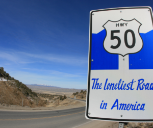 Highway 50 road sign reading “The Loneliest Road in America” along a rural desert highway under a wide blue sky.