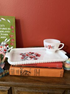 Vintage floral teacup resting on a white milk glass tray atop stacked antique Alice in Wonderland books, arranged on a wooden table against a warm red wall, creating a cozy, bookish tea vignette.