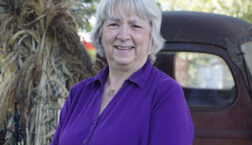 Author Jenny Carlisle smiling outdoors in a purple blouse, standing beside a vintage truck and fall decorations.