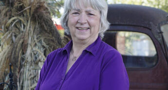 Author Jenny Carlisle smiling outdoors in a purple blouse, standing beside a vintage truck and fall decorations.