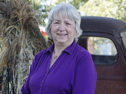 Author Jenny Carlisle smiling outdoors in a purple blouse, standing beside a vintage truck and fall decorations.