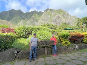Couple standing by a sign at Kualoa Ranch in Hawaii with lush green mountains and tropical landscape behind them.