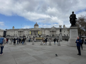 Visitors walking through Trafalgar Square in London with the National Gallery and statues visible in the background.