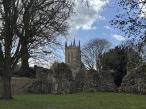 Ancient stone ruins in a grassy area with a cathedral tower rising in the background under a partly cloudy sky.