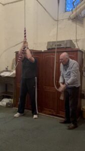 Two people ringing church bells inside a historic tower, demonstrating traditional bell-ringing techniques with ropes.