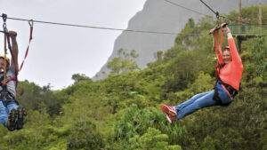 Woman ziplining through a lush tropical landscape with green mountains and trees in the background.