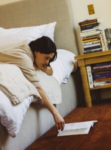 Woman lying on bed reaching for a book on the floor beside a nightstand stacked with books.